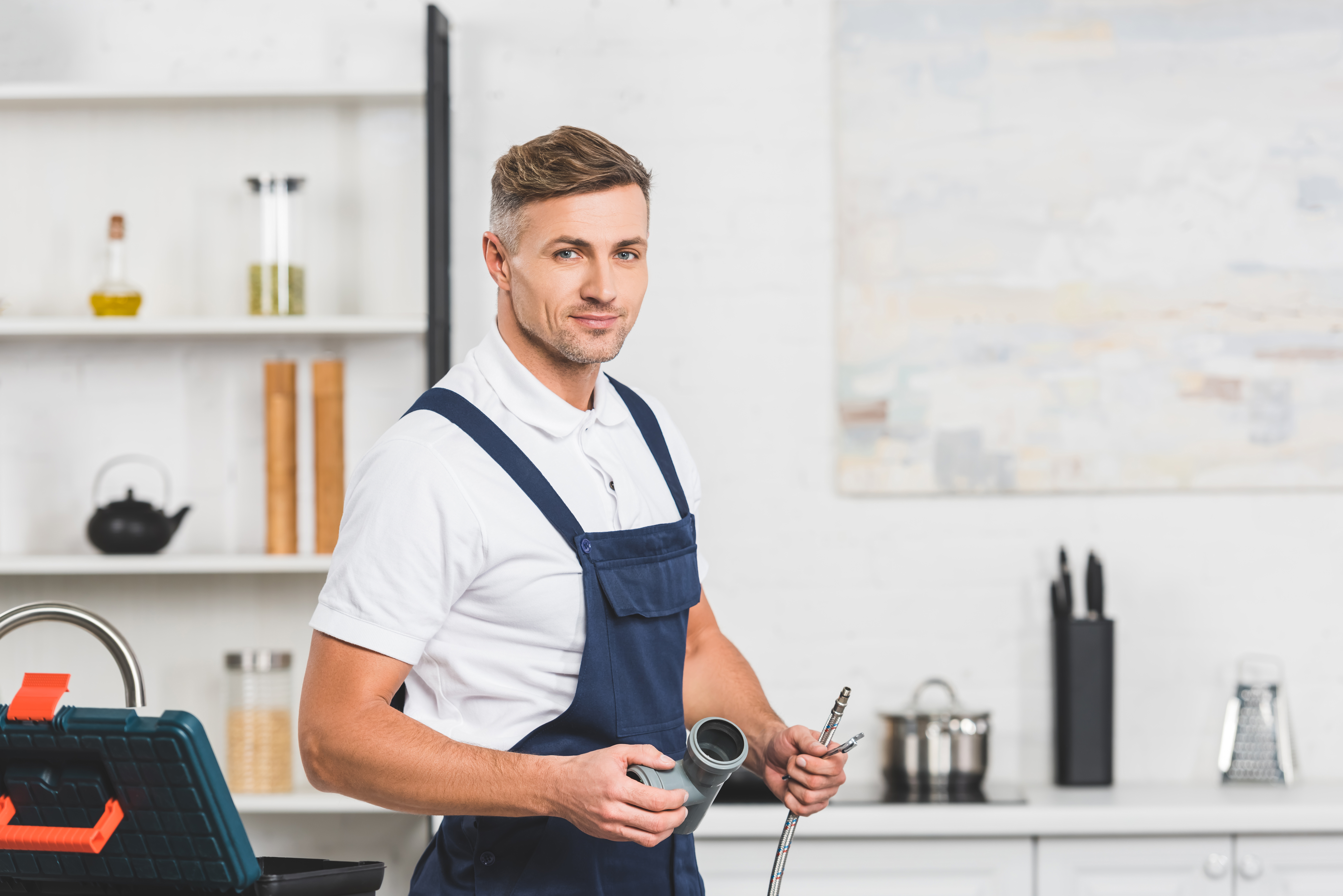 A Man holding plumbing items with his tool box open in a modern white looking kitchen.  A shelf and sink are behind him with multiple kitchen items