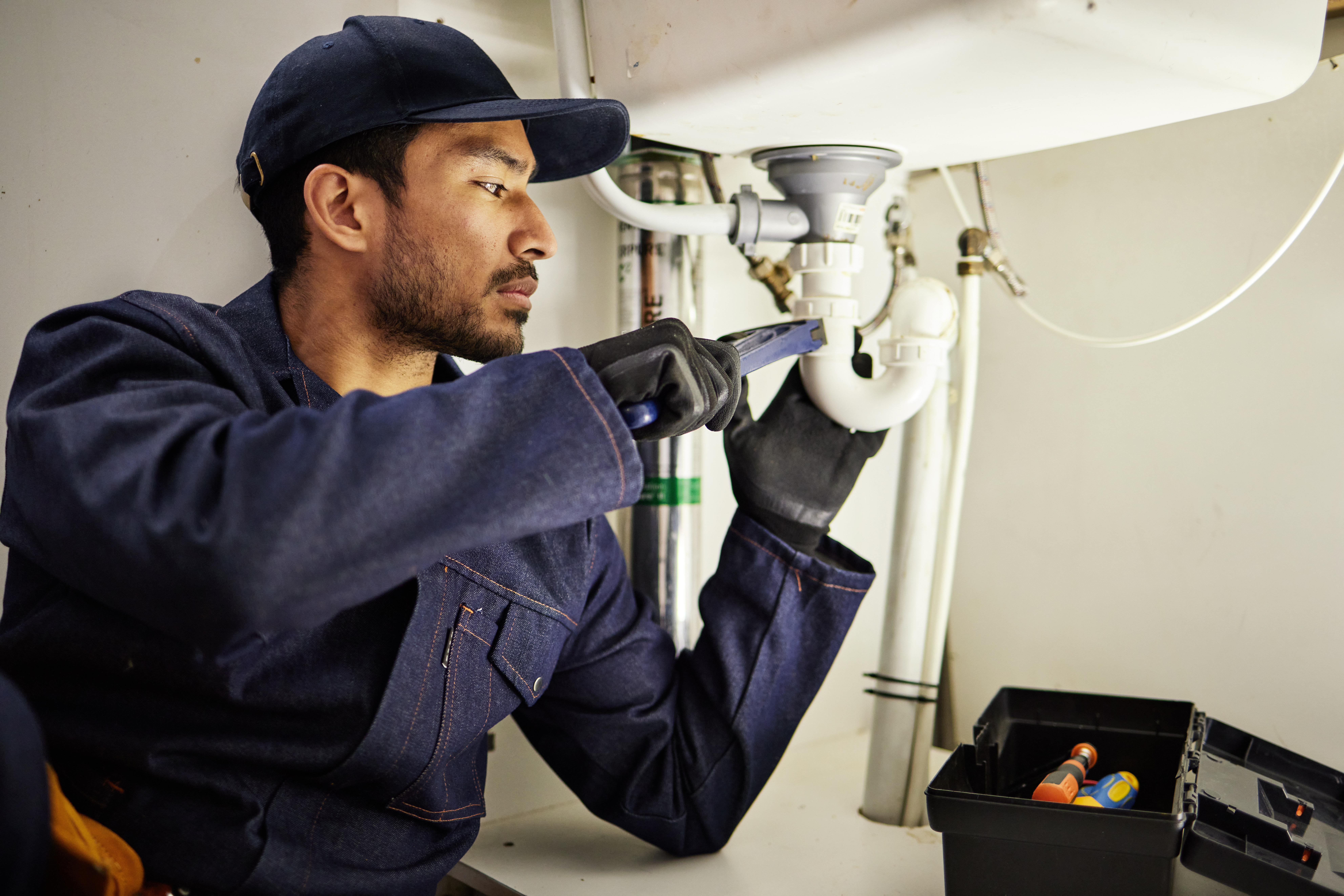 Plumber under a sink wearing a blue plumber suite.  Tightening a fitting attached to a sink in a white bathroom.