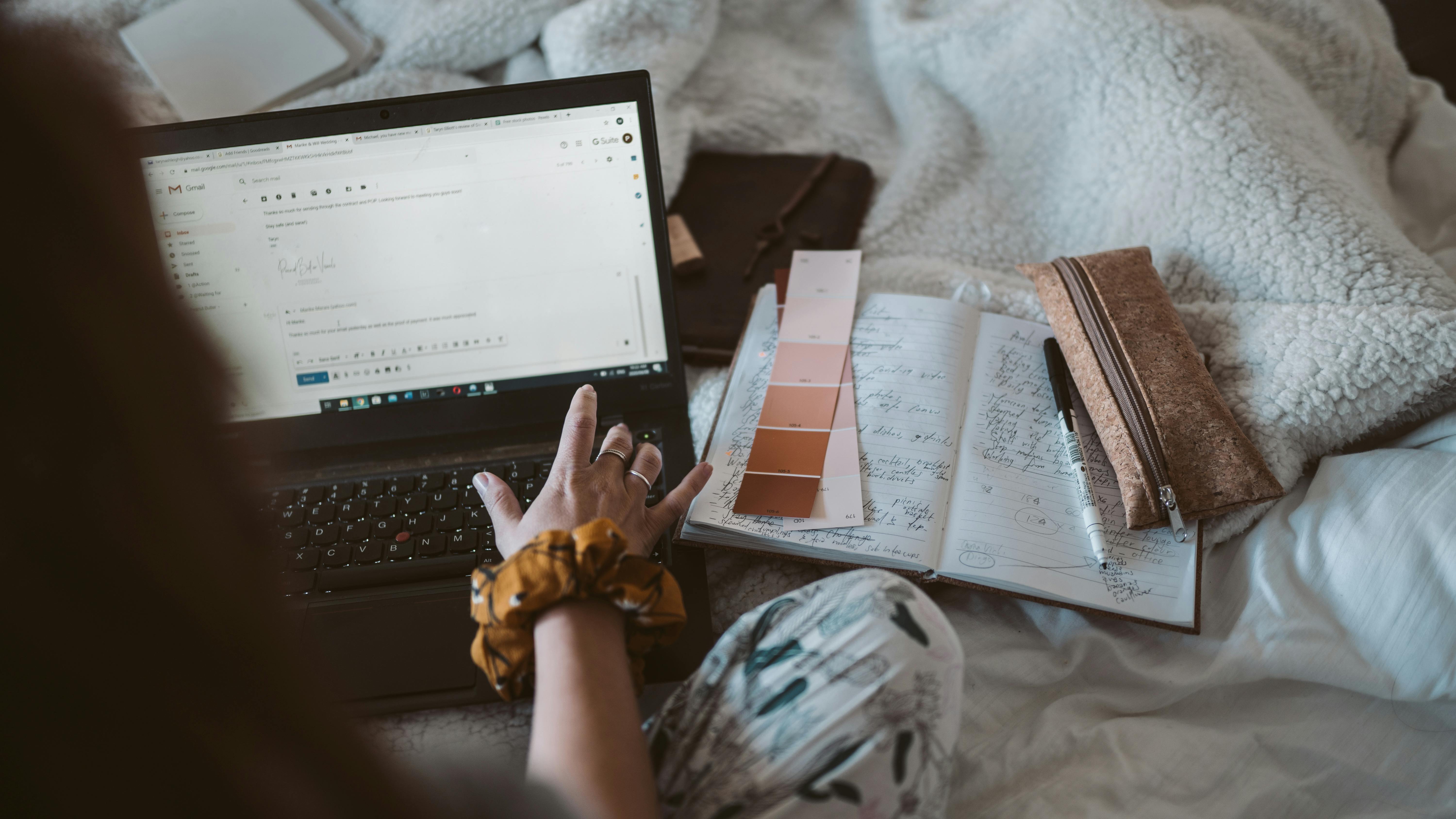 Woman emailing on a laptop on a cozy bed with color swatches to her side and a open notebook.  A wallet and pen lay on top of the notebook to the right.