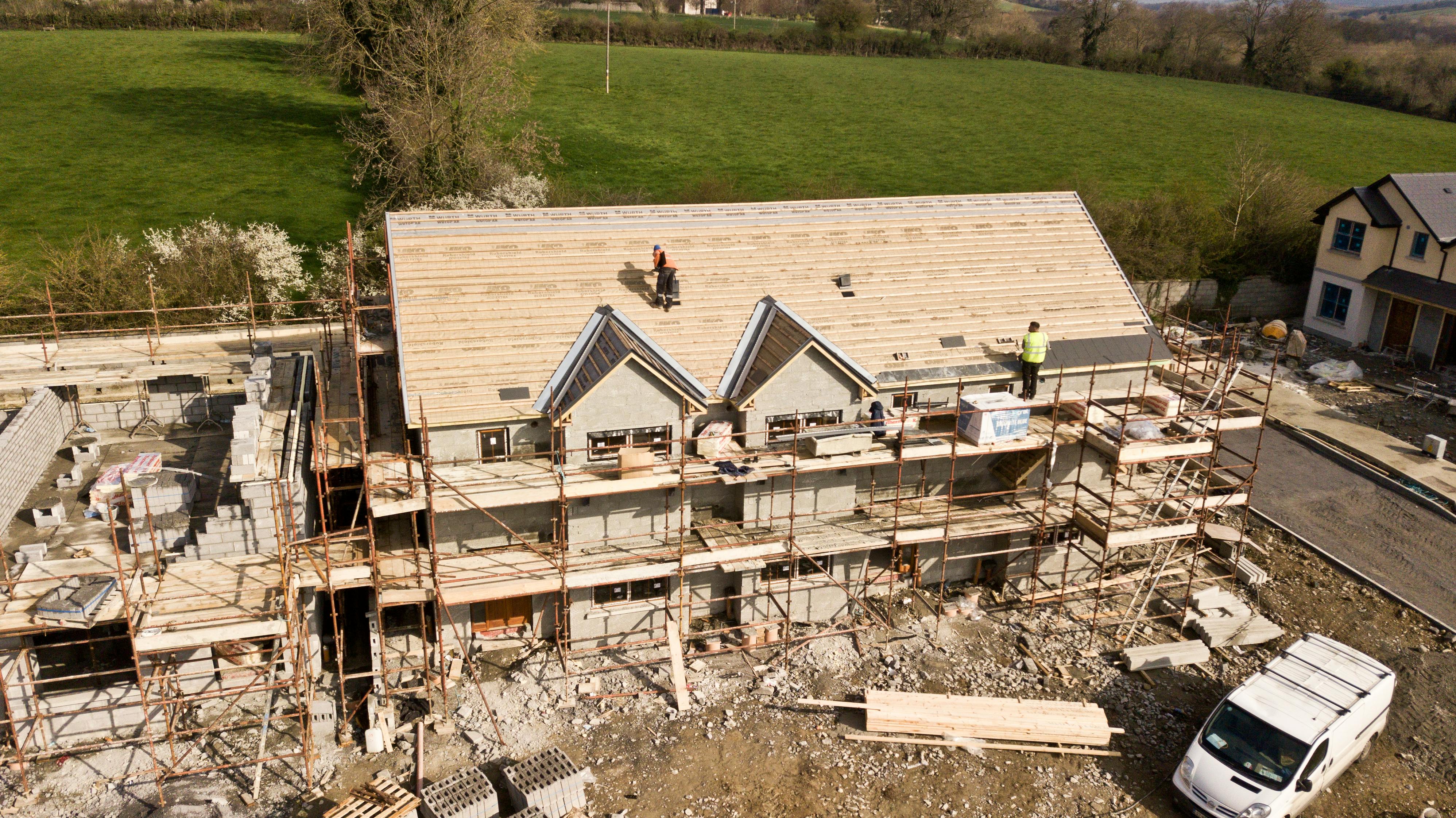 The image shows a construction site of what appears to be a residential building in its framing phase. There are exposed wooden roof trusses on the top of the structure, and a crew can be seen working on the roof, which is partially covered with sheathing. The house is a two-story structure with visible masonry work on the first floor. There's scaffolding around the building, indicating ongoing work. Construction materials and tools are scattered across the site. A white van is parked in the foreground, and a lush green field lies in the background under a clear sky, suggesting a rural or suburban setting.
